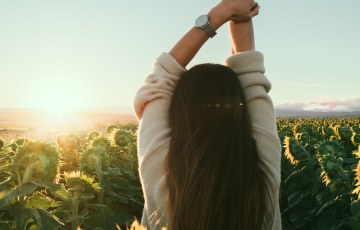 Nainen seisoo selin kameraan auringonkukkapellossa auringon laskiessa