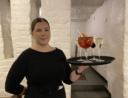 Tarjoilijaa pitelee tarjotinta, jossa on täysiä valkoviinilaseja sekä yksi iso drinkki. Taustalla Kellariravintola Hällän tiiliseinää. 